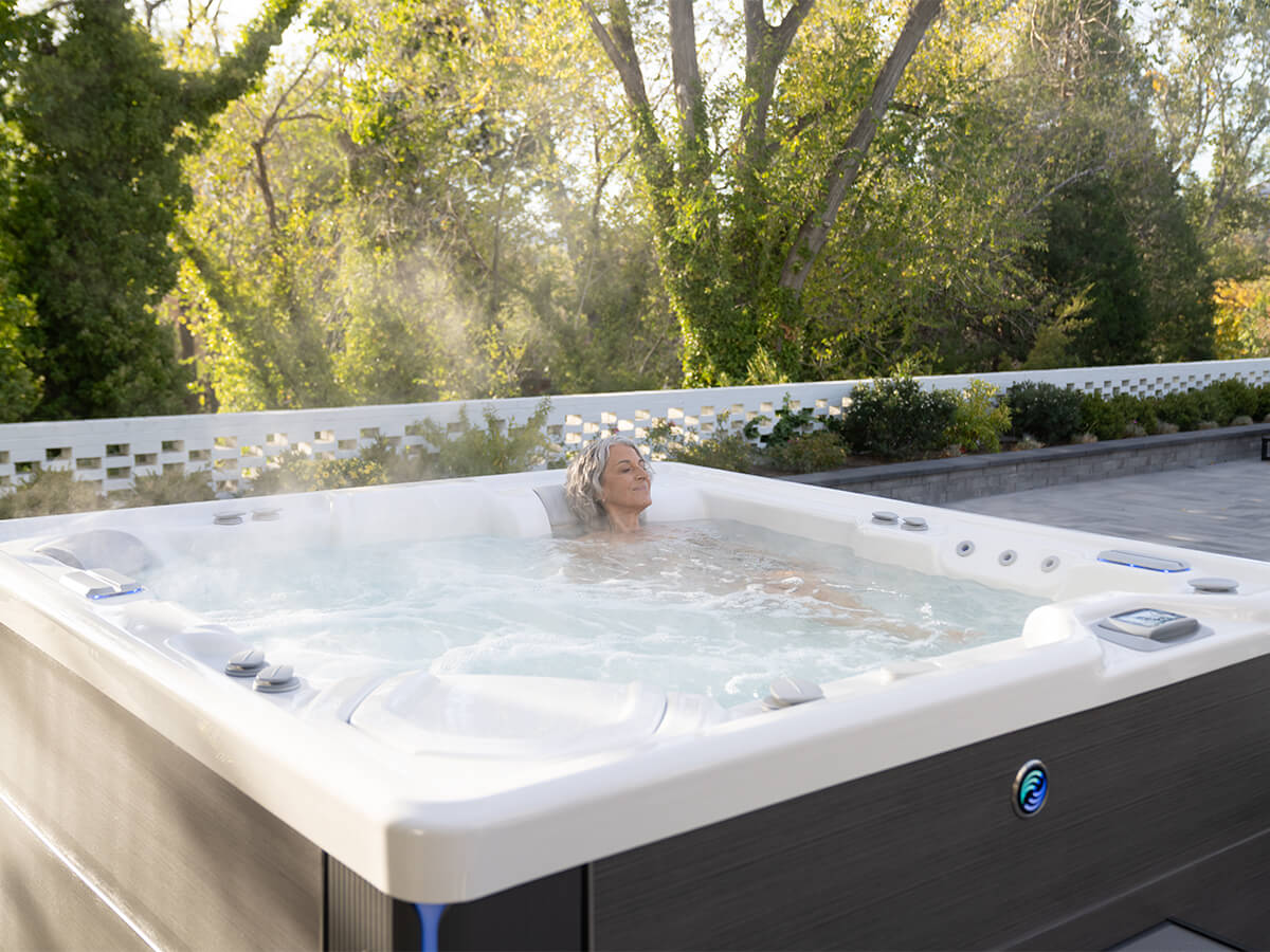 A woman is enjoying a soak in her Hot Spring Spas hot tub in an outdoor backyard setting. The hot tub is surrounded by lush greenery. The woman is relaxed and content, enjoying the peace and quiet of her backyard.