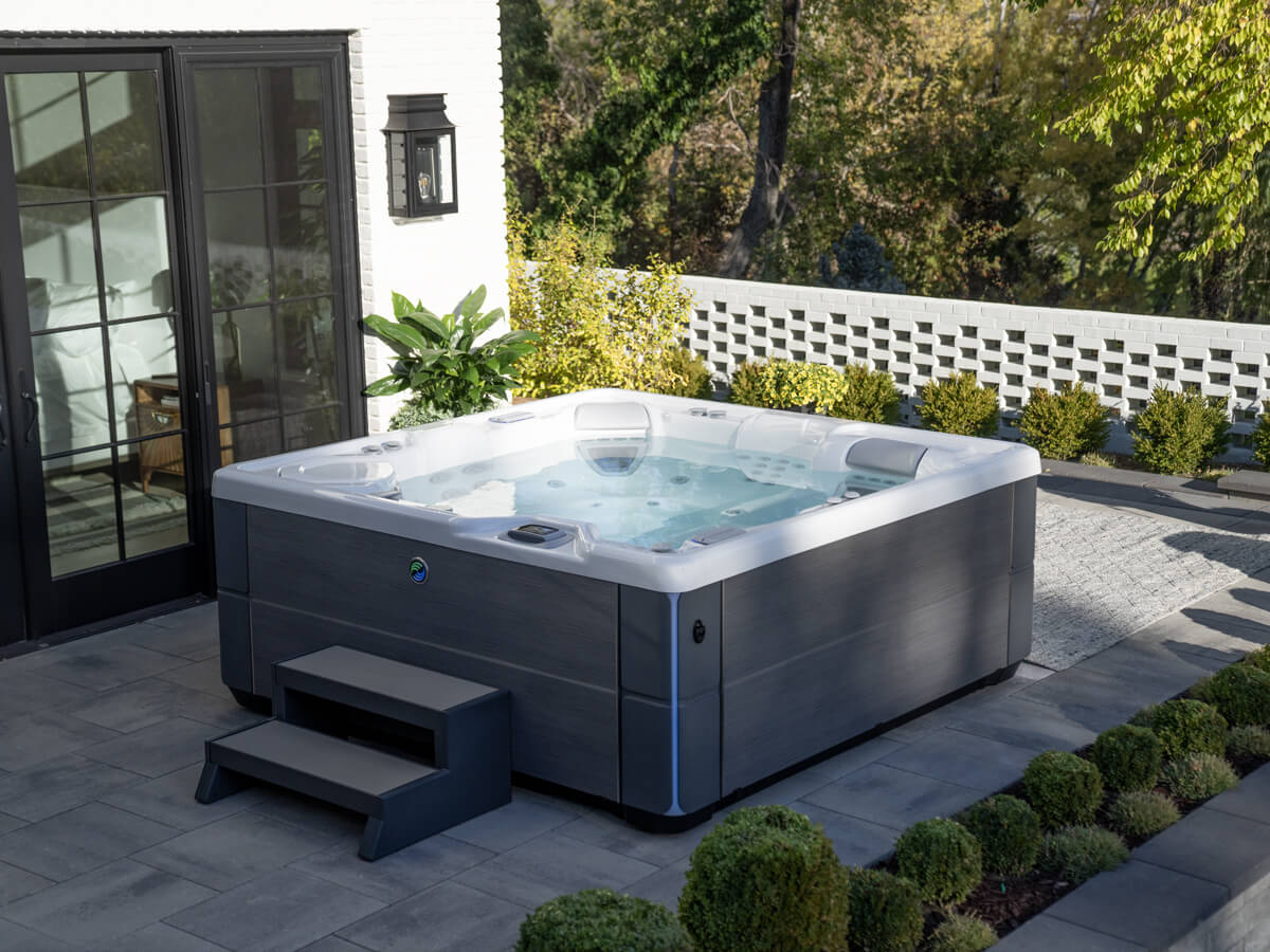 A Highlife Aria salt water hot tub sits on a stone patio with easy access provided by side steps. The hot tub is surrounded by plants.