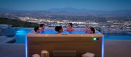A group of six people relaxes in a Hot Spring Spas hot tub on a patio overlooking a city at dusk.