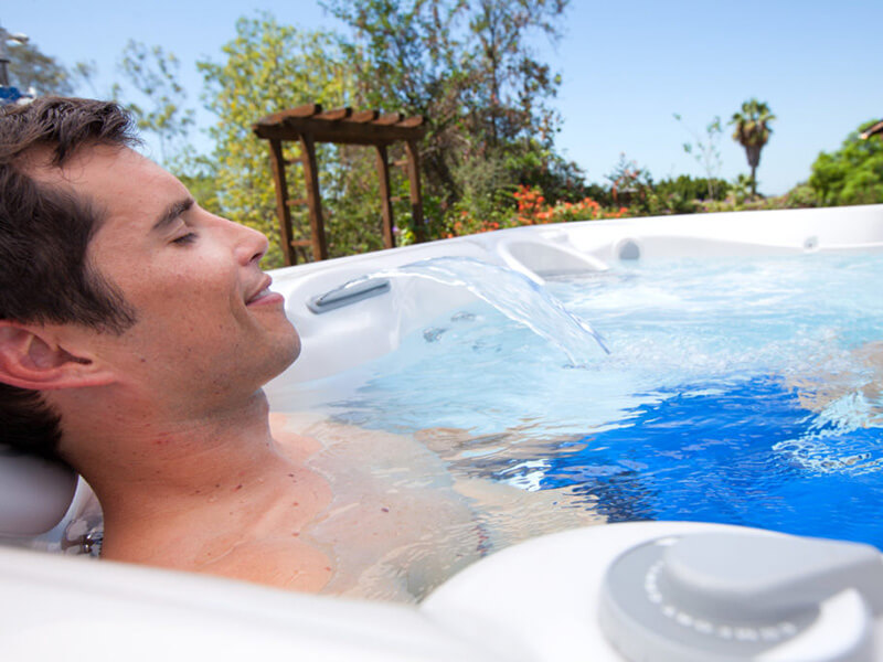 Man relaxing in an outdoor hot tub with a soothing waterfall feature, surrounded by lush greenery and a pergola in the background.