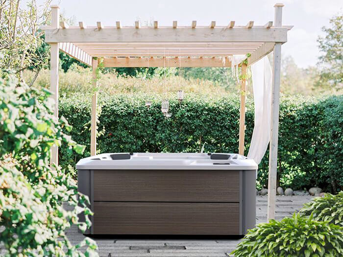 Elegant outdoor hot tub placed under a wooden pergola with hanging lanterns and flowing white drapes, surrounded by lush greenery.