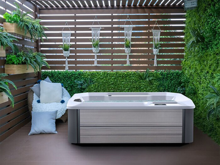 Modern outdoor hot tub in a serene, plant-filled space with a wooden privacy screen, hanging planters, and a cozy seating area with plush pillows.