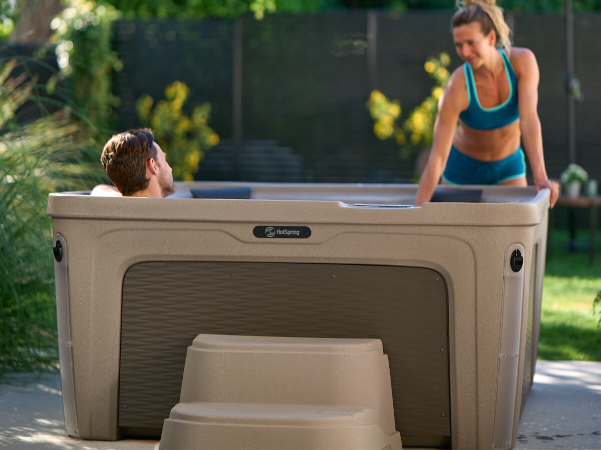 A couple enjoying a Hot Spring plug-and-play hot tub together in a lush backyard garden, with one person soaking inside and another leaning over the edge