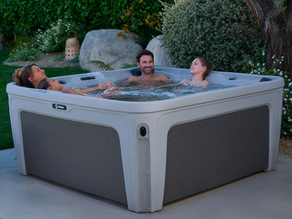 A family of three — two adults and a child — laughing and playing together in a rotomolded Hot Spring hot tub in a landscaped backyard