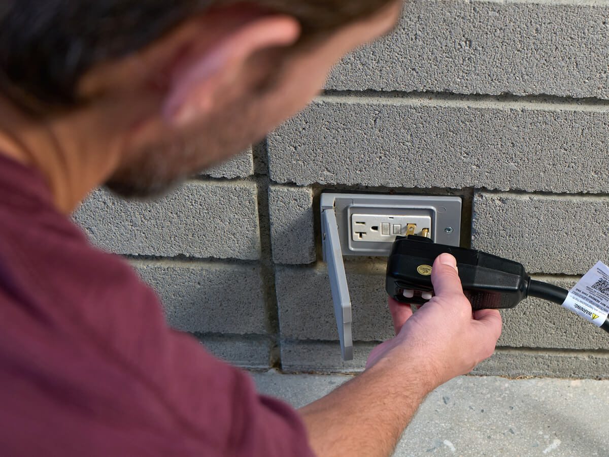 A man plugging a power cord into an outdoor electrical outlet mounted on a brick wall
