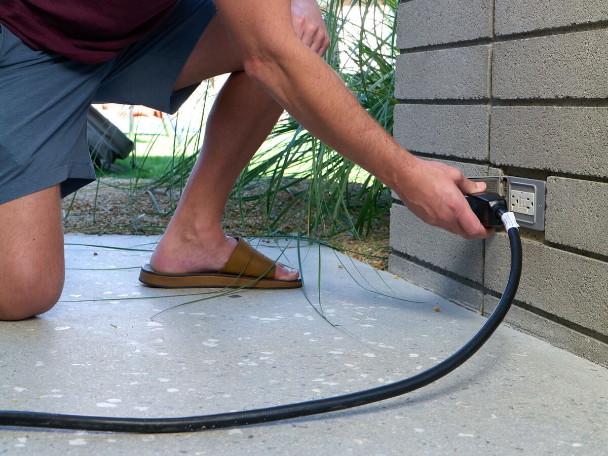 A person kneeling on a concrete patio plugging a power cord into an outdoor electrical outlet mounted on a wall